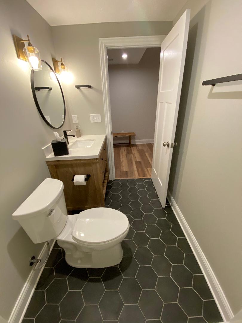 Updated bathroom with round black mirror, brass sconces, wood vanity and hex tile floor